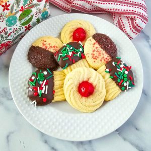 A festive display of Christmas butter cookies, featuring a variety of decorative styles. Some cookies are dipped in chocolate and adorned with colorful sprinkles, while others are garnished with cherries or red sugar crystals. The cookies are arranged on a white plate and in a gift box, surrounded by a red and white striped fabric for a cozy holiday vibe.