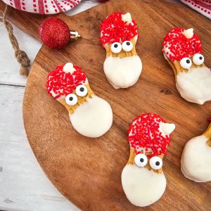 A festive platter of homemade Santa Nutter Butter cookies on a wooden board. Each cookie is decorated with white chocolate for a beard and red sprinkles for a hat.