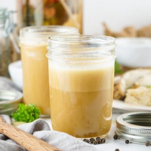 Two mason jars filled with homemade chicken stock sit on a white surface. A plate of food and a wooden spoon are also visible.