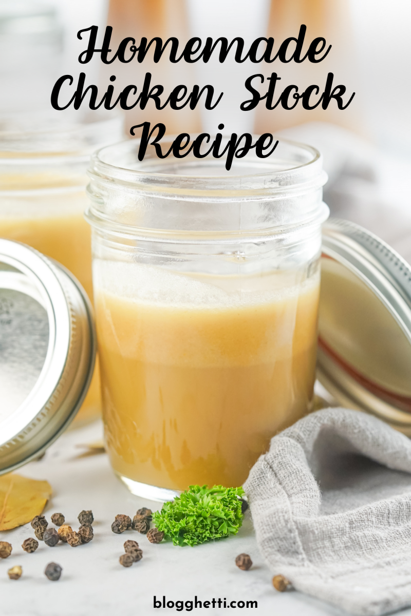 A mason jar filled with golden chicken stock sits on a white surface. Black peppercorns, bay leaves, and parsley are scattered around the jar.