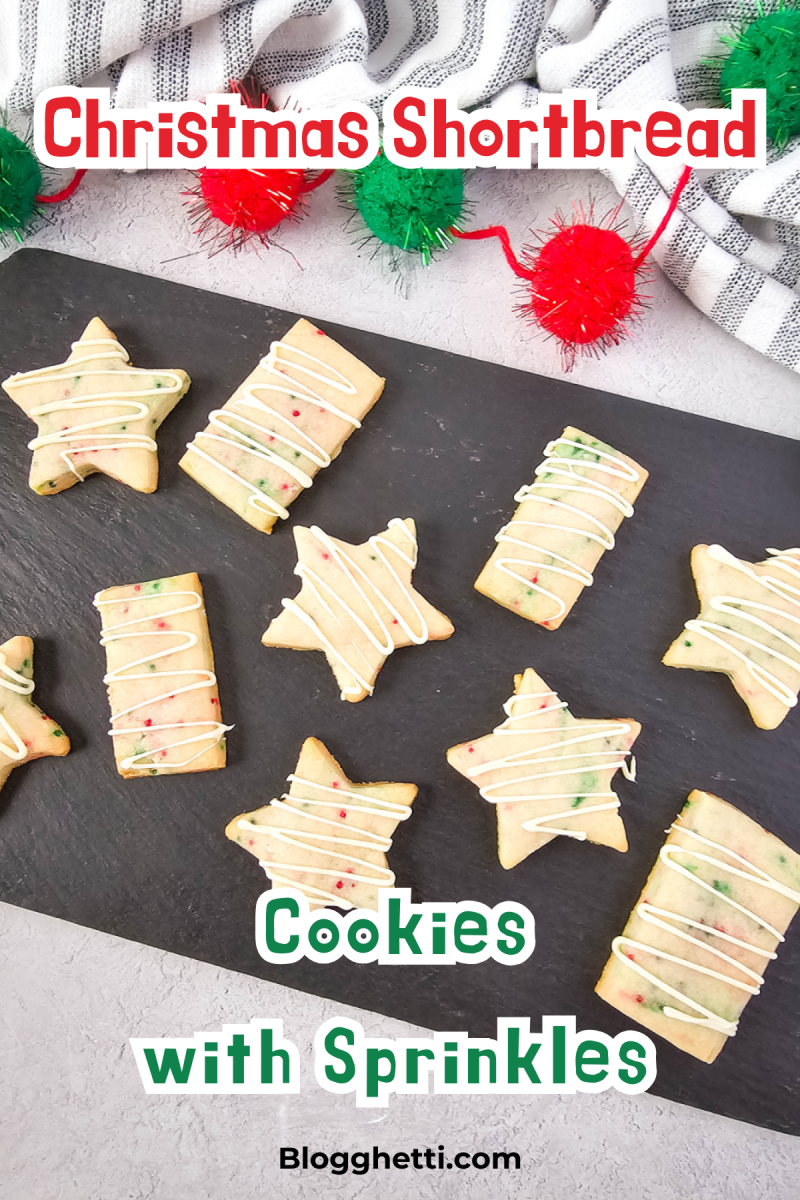 A top-down view of a black serving board with an assortment of festive Christmas shortbread cookies. The cookies are cut into star and rectangle shapes and are decorated with colorful sprinkles and white icing. Red and green pom-poms and a striped towel are in the background.