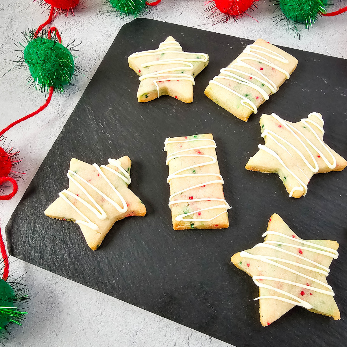 A top-down view of a black serving board with an assortment of festive Christmas shortbread cookies. The cookies are cut into star and rectangle shapes and are decorated with colorful sprinkles and white icing. Red and green pom-poms are in the background.