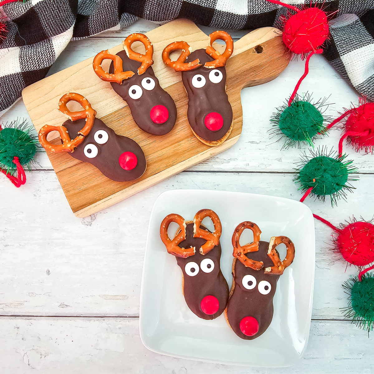 A close-up image of three Nutter Butter Reindeer Cookies on a wooden cutting board. Each cookie has chocolate frosting, candy eyes, a red nose, and pretzel antlers.