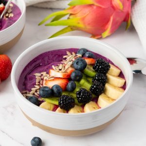 A vibrant purple dragon fruit smoothie bowl topped with sliced banana, kiwi, blackberries, blueberries, strawberries, and pumpkin seeds