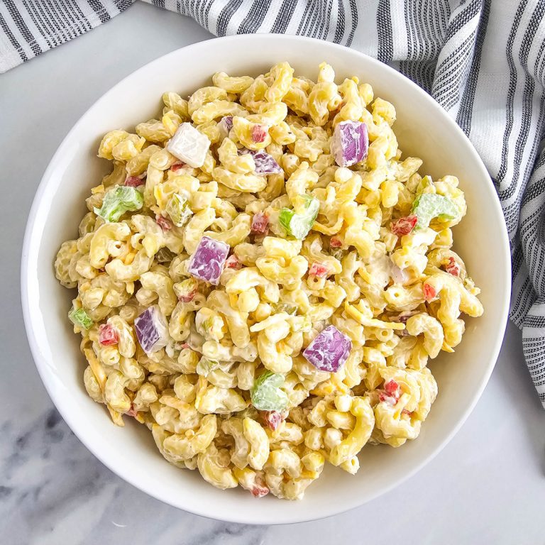 A bowl of creamy macaroni salad, viewed from above. Elbow macaroni is mixed with mayonnaise dressing, red onion, celery, cheese, and pimentos. A striped kitchen towel lies on the light surface next to the white bowl.
