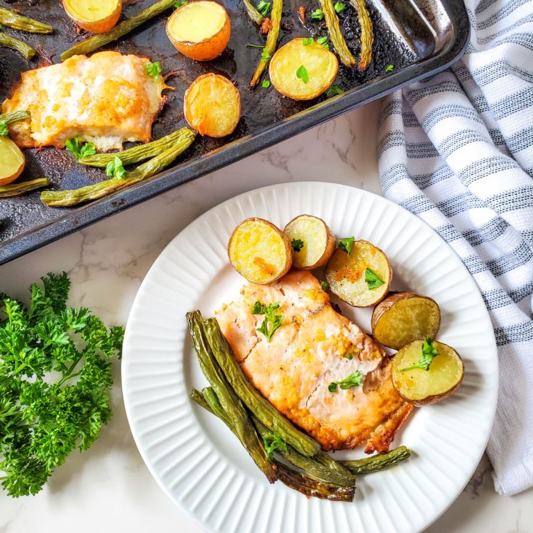 Overhead shot of a white plate with a serving of baked salmon, roasted halved potatoes, and cooked green beans, garnished with fresh parsley. A baking sheet with more salmon, potatoes, and green beans is visible in the upper left corner. A bunch of fresh parsley sits on the white marble surface next to the plate.