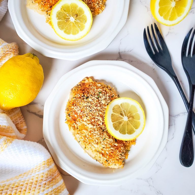 Overhead shot of two white plates each holding a parmesan-crusted fish fillet, likely tilapia, garnished with a lemon slice. Black forks are placed on the white marble countertop beside the plates. A whole lemon and a folded yellow and white striped kitchen towel are in the upper left corner of the frame.