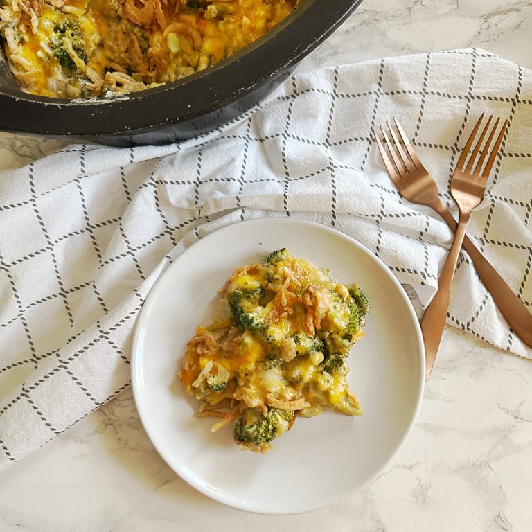A serving of cheesy broccoli casserole with crispy fried onions sits on a white plate, with the remaining casserole in a black slow cooker in the background. The dish is placed on a white and black checkered kitchen towel, accompanied by two copper forks on a marble countertop.