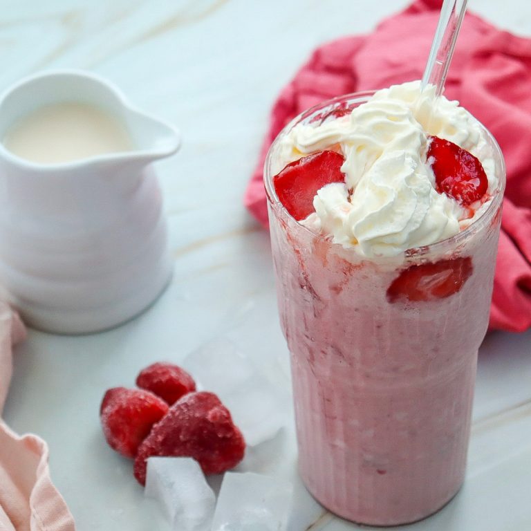 A pink Strawberry Cheesecake Frappuccino in a clear glass sits on a white marble surface, topped with swirls of white whipped cream and slices of bright red strawberries. A clear straw is inserted into the drink. To the left are three frozen strawberry halves and several clear ice cubes.