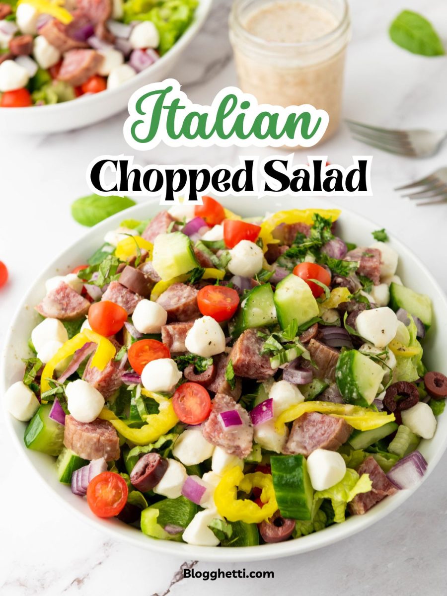 A vibrant Italian Chopped Salad in a white bowl features chopped Romaine lettuce, halved cherry tomatoes, diced cucumber, red onion, black olives, cubed fresh mozzarella, diced green and yellow bell peppers, sliced salami, and fresh herbs. A jar of creamy dressing and another salad bowl are visible in the blurred background on a white marble surface.