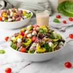 A vibrant Italian Chopped Salad in a white bowl features chopped Romaine lettuce, halved cherry tomatoes, diced cucumber, red onion, black olives, cubed fresh mozzarella, diced peppers, sliced salami, and fresh herbs. A jar of creamy dressing and another salad bowl are visible in the blurred background on a white marble surface.