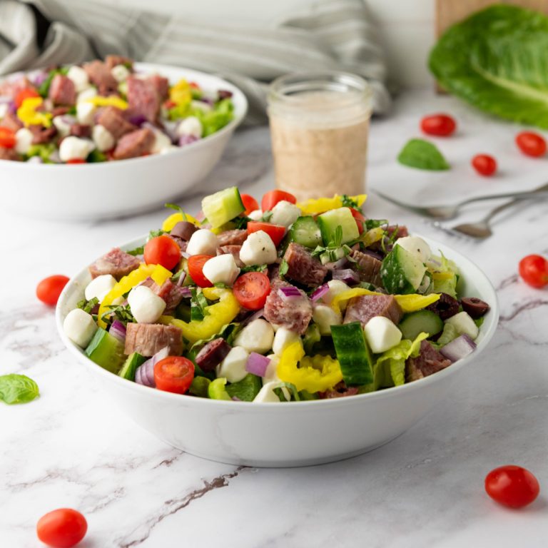 A vibrant Italian Chopped Salad in a white bowl features chopped Romaine lettuce, halved cherry tomatoes, diced cucumber, red onion, black olives, cubed fresh mozzarella, diced peppers, sliced salami, and fresh herbs. A jar of creamy dressing and another salad bowl are visible in the blurred background on a white marble surface.