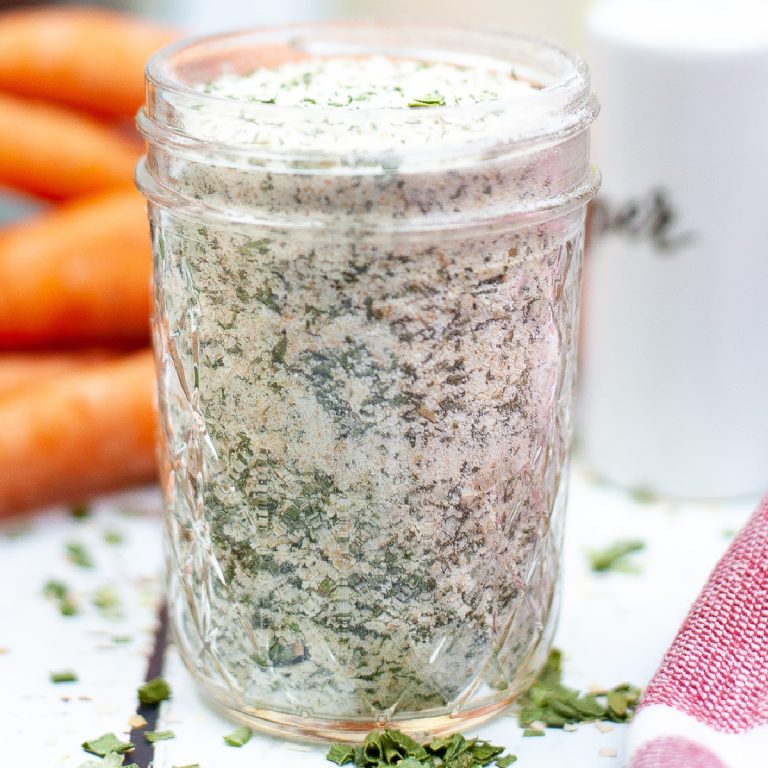 Close-up of a glass jar filled with dry homemade ranch seasoning mix, with carrots and a white shaker in the blurred background.