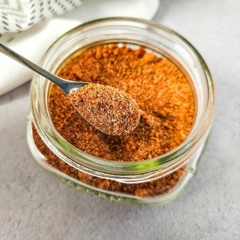 Close-up image of a spoonful of homemade burger seasoning held above a glass jar filled with the spice blend. The seasoning is a mix of finely ground spices in shades of red and brown, with a soft background and white kitchen towel partially visible.