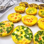 A close-up shot shows a white rectangular plate filled with twelve individual muffin tin omelets. The omelets are round, golden-yellow, and showcase various colorful fillings, including green onions, red bell peppers, and spinach. With some having bacon and sausage. A white and black checkered kitchen towel is visible in the upper left corner of the frame, slightly blurred. The lighting is bright, highlighting the texture and vibrant colors of the mini omelets.
