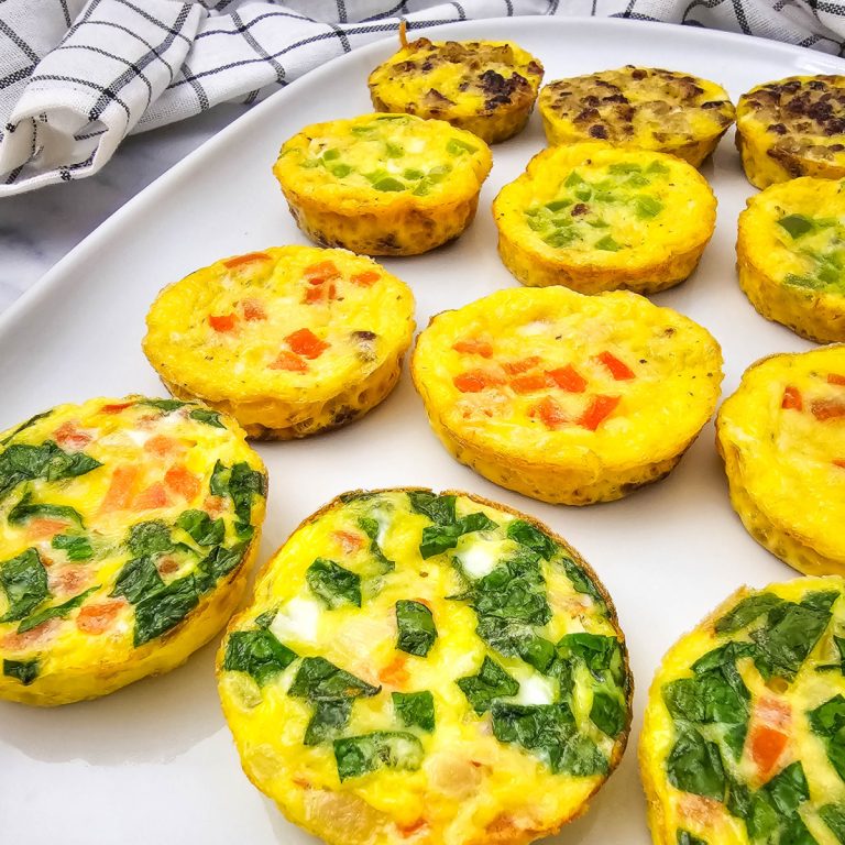 A close-up shot shows a white rectangular plate filled with twelve individual muffin tin omelets. The omelets are round, golden-yellow, and showcase various colorful fillings, including green onions, red bell peppers, and spinach. With some having bacon and sausage. A white and black checkered kitchen towel is visible in the upper left corner of the frame, slightly blurred. The lighting is bright, highlighting the texture and vibrant colors of the mini omelets.