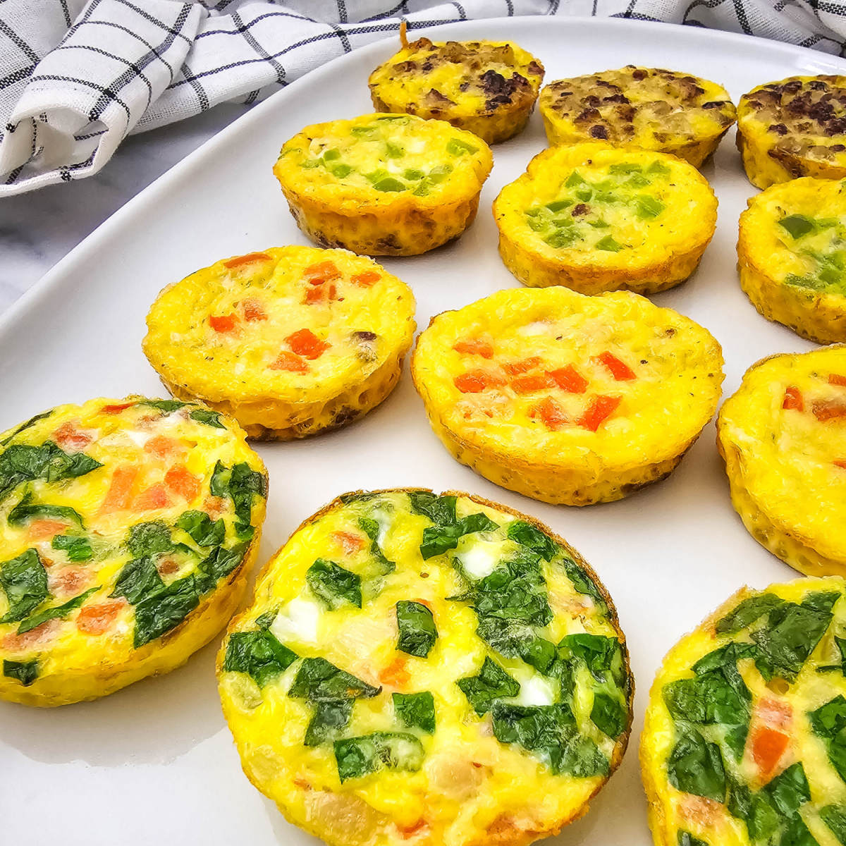 A close-up shot shows a white rectangular plate filled with twelve individual muffin tin omelets. The omelets are round, golden-yellow, and showcase various colorful fillings, including green onions, red bell peppers, and spinach. With some having bacon and sausage. A white and black checkered kitchen towel is visible in the upper left corner of the frame, slightly blurred. The lighting is bright, highlighting the texture and vibrant colors of the mini omelets.