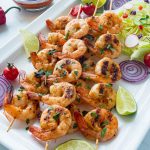 A rectangular white platter holds several grilled Chipotle Lime Shrimp skewers, garnished with fresh lime wedges and chopped cilantro. The platter is also decorated with a small pile of lettuce, sliced red onions, cherry tomatoes, and radish slices. A small glass bowl containing a reddish sauce with a spoon rests near the top of the platter. In the background, on a white marble surface, are a bottle of oil, a whole red pepper, a halved lime, and a sprig of parsley.