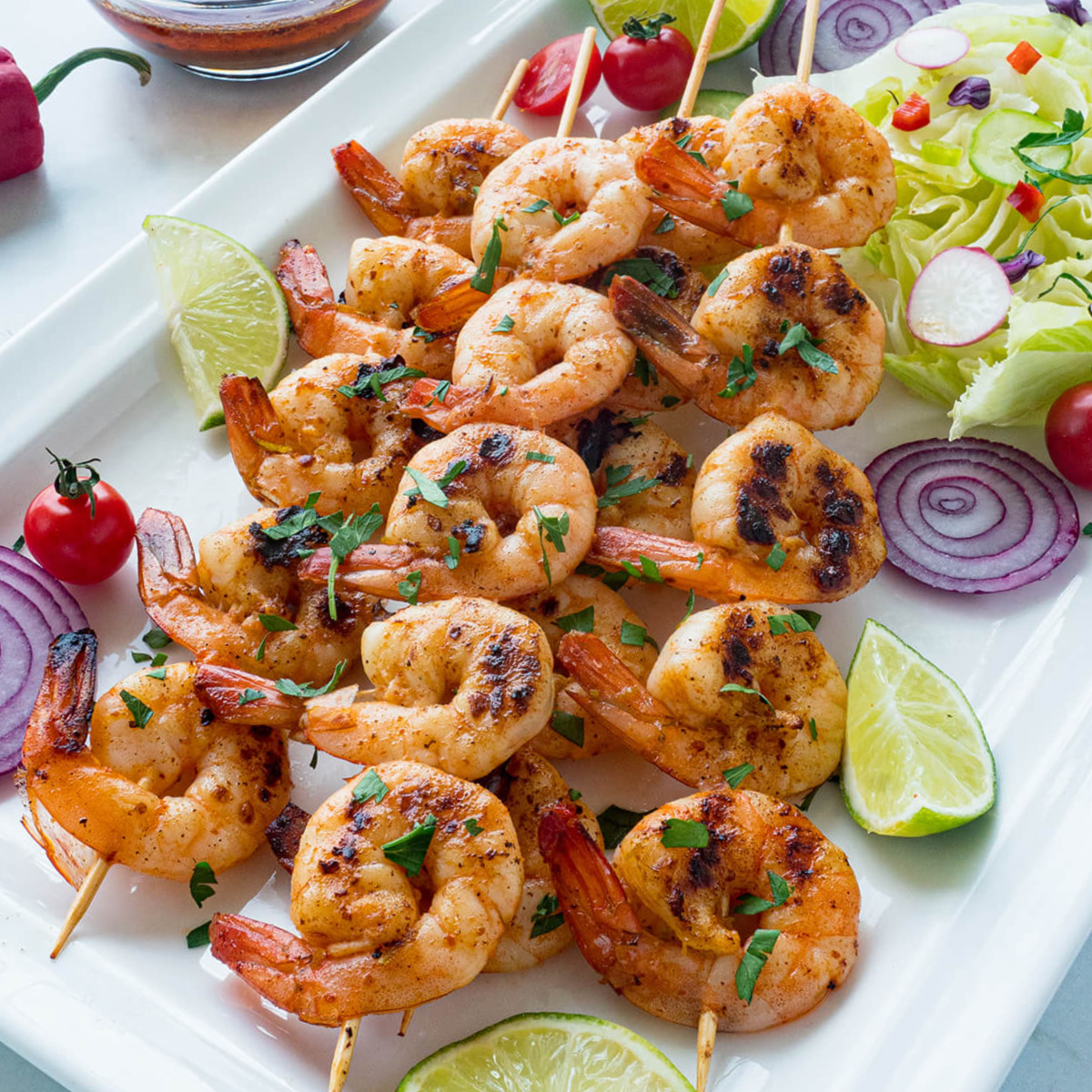 A rectangular white platter holds several grilled Chipotle Lime Shrimp skewers, garnished with fresh lime wedges and chopped cilantro. The platter is also decorated with a small pile of lettuce, sliced red onions, cherry tomatoes, and radish slices. A small glass bowl containing a reddish sauce with a spoon rests near the top of the platter. In the background, on a white marble surface, are a bottle of oil, a whole red pepper, a halved lime, and a sprig of parsley.