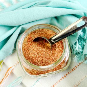 Overhead close-up of a clear glass jar filled with reddish-brown BBQ dry rub seasoning. A silver measuring spoon with visible measurement markings rests inside the jar, partially scooping the spice blend. The jar sits on a white surface with subtle colored stripes and fringe, partially covered by a soft, light blue cloth.