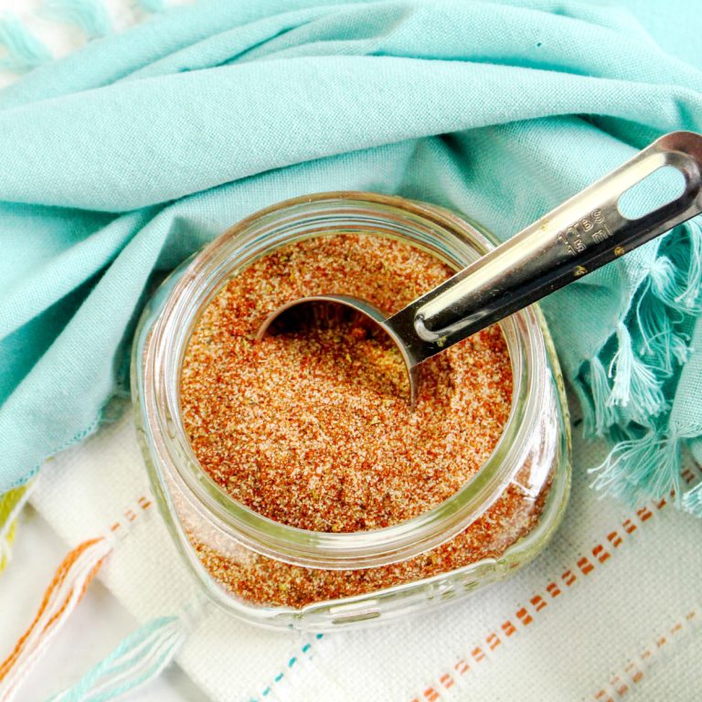 Overhead close-up of a clear glass jar filled with reddish-brown BBQ dry rub seasoning. A silver measuring spoon with visible measurement markings rests inside the jar, partially scooping the spice blend. The jar sits on a white surface with subtle colored stripes and fringe, partially covered by a soft, light blue cloth.