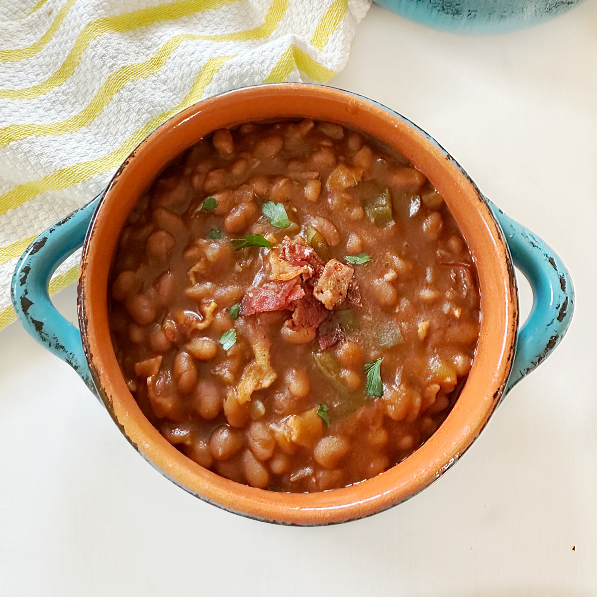 A close-up of baked beans served in an orange bowl with blue handles. Crispy bacon pieces are scattered on top, along with a fresh parsley garnish. A white and yellow striped towel is partially visible in the background, adding a warm, rustic touch to the scene.