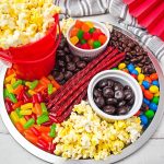 A silver round charcuterie board filled with movie night snacks including red buckets of popcorn, colorful gummy candies, red licorice sticks, chocolate-covered raisins, and M&M's.