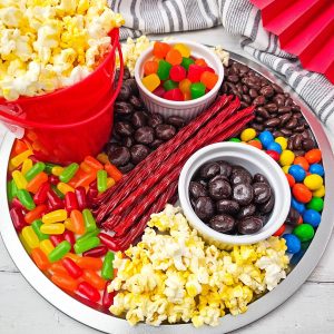 A silver round charcuterie board filled with movie night snacks including red buckets of popcorn, colorful gummy candies, red licorice sticks, chocolate-covered raisins, and M&M's.