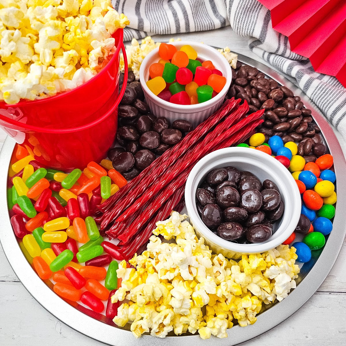 A silver round charcuterie board filled with movie night snacks including red buckets of popcorn, colorful gummy candies, red licorice sticks, chocolate-covered raisins, and M&M's.