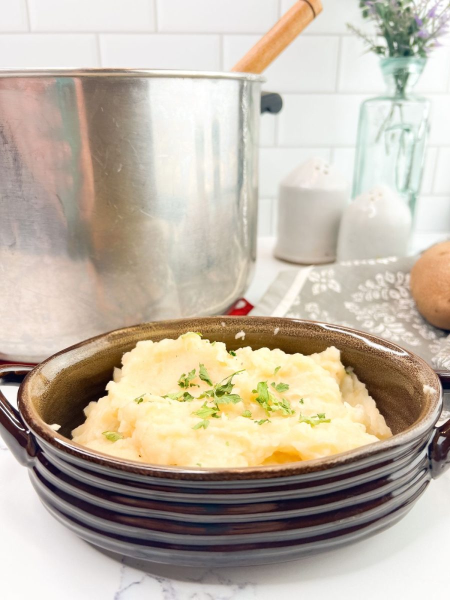 A close-up shot of a small, dark brown ceramic bowl overflowing with fluffy mashed potatoes, topped with chopped green parsley, sitting on a white marble countertop. In the background, a large metal pot with a wooden handle, salt and pepper shakers, a patterned grey kitchen towel, and a vase with purple flowers are visible.