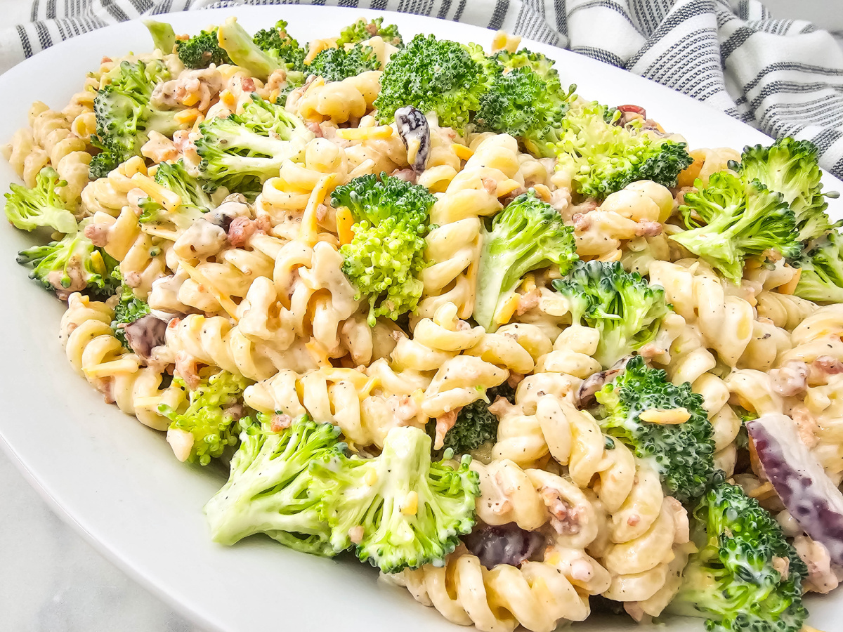 A wide white oval serving platter holds a large portion of creamy broccoli pasta salad, featuring rotini pasta, bright green broccoli florets, cheese, and red onion pieces, with a striped napkin in the background.