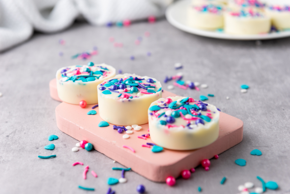 Three round white chocolate covered Oreo cookies, each adorned with colorful sprinkles, are displayed on a pink cutting board. More cookies are out of focus on a white plate in the background