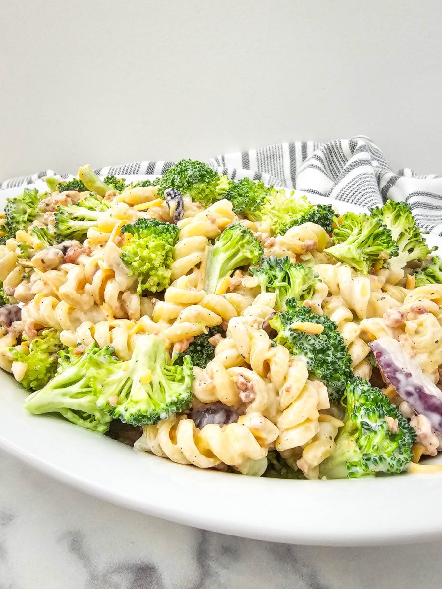 A close-up, slightly elevated view of creamy broccoli pasta salad on a white oval platter, showing detailed textures of rotini pasta, broccoli, and a creamy dressing, with a patterned cloth behind.