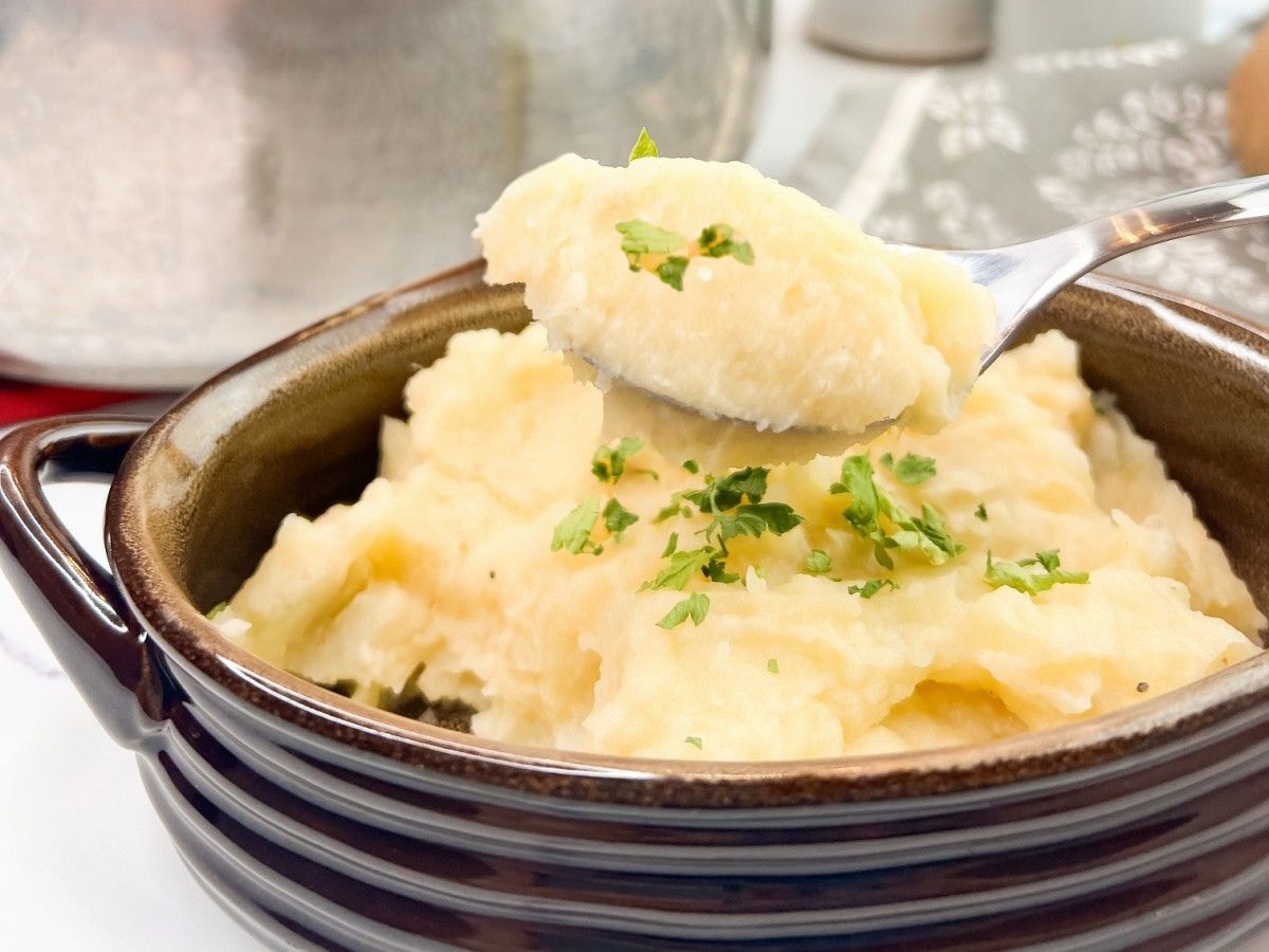 A close-up shot of a silver spoon lifting a perfect scoop of creamy mashed potatoes from a brown ceramic bowl. The potatoes are light yellow and appear smooth, with specks of green parsley scattered on top and in the bowl.