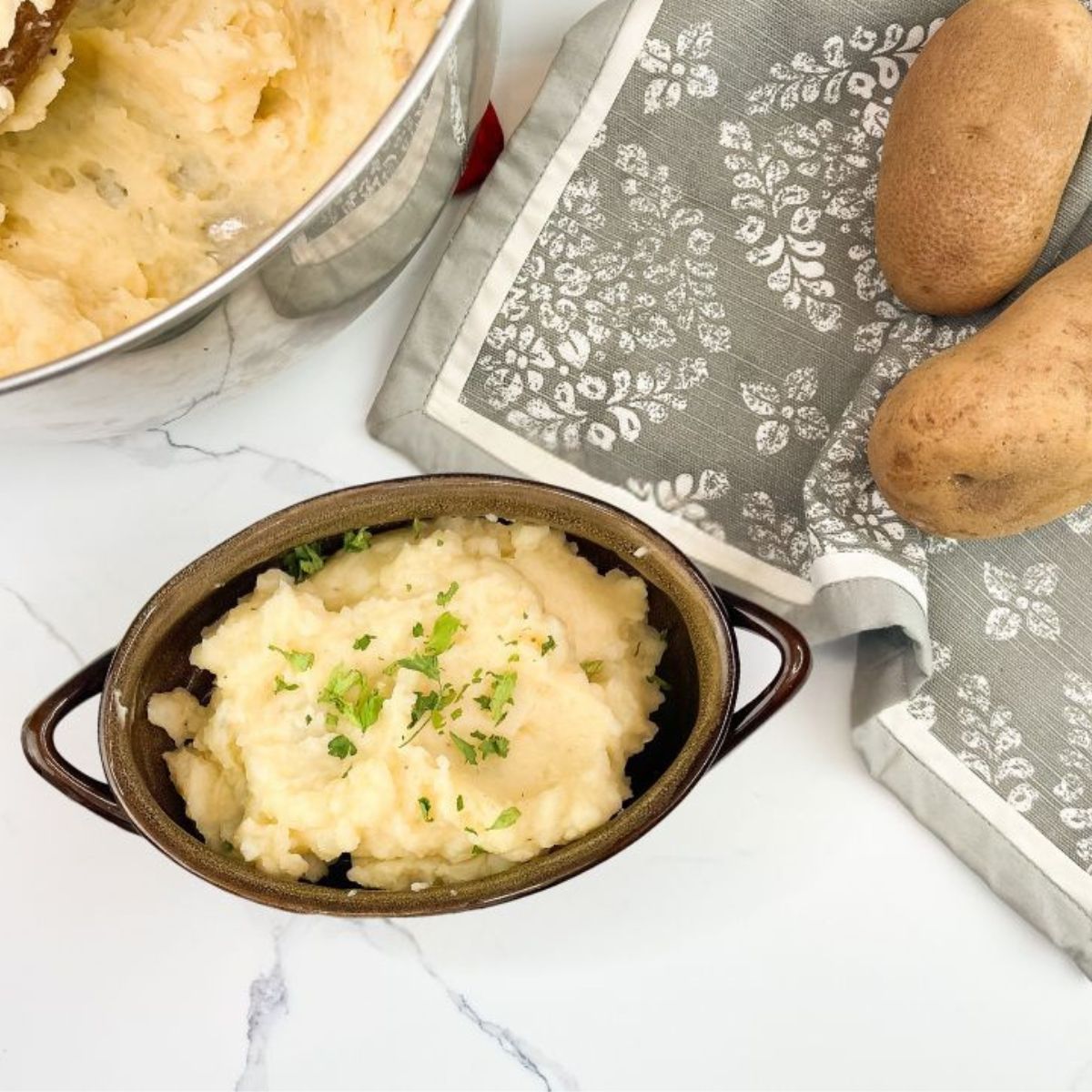An overhead shot of a small brown bowl filled with creamy mashed potatoes garnished with parsley. Next to it, a patterned grey kitchen towel lies on a white marble surface with two whole russet potatoes. In the upper left, a large metal mixing bowl containing more mashed potatoes is partially visible.