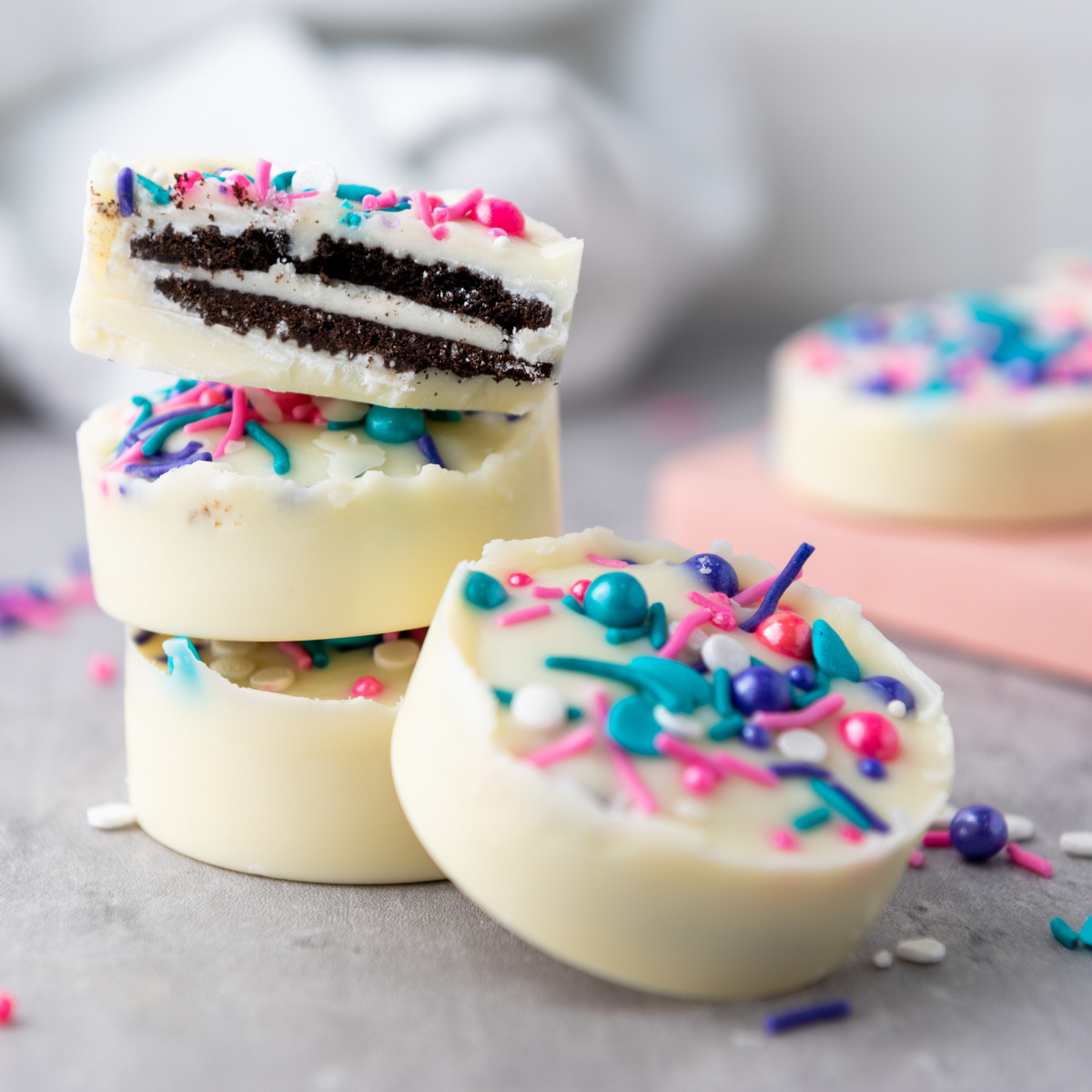 A stack of two white chocolate covered Oreos with colorful sprinkles, with the top cookie cut in half revealing the black Oreo cookie inside. Another full white chocolate covered Oreo rests in front of the stack.