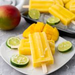 Several bright yellow mango coconut lime popsicles with wooden sticks are arranged on a white plate with lime slices, shredded coconut, and a whole mango in the background.
