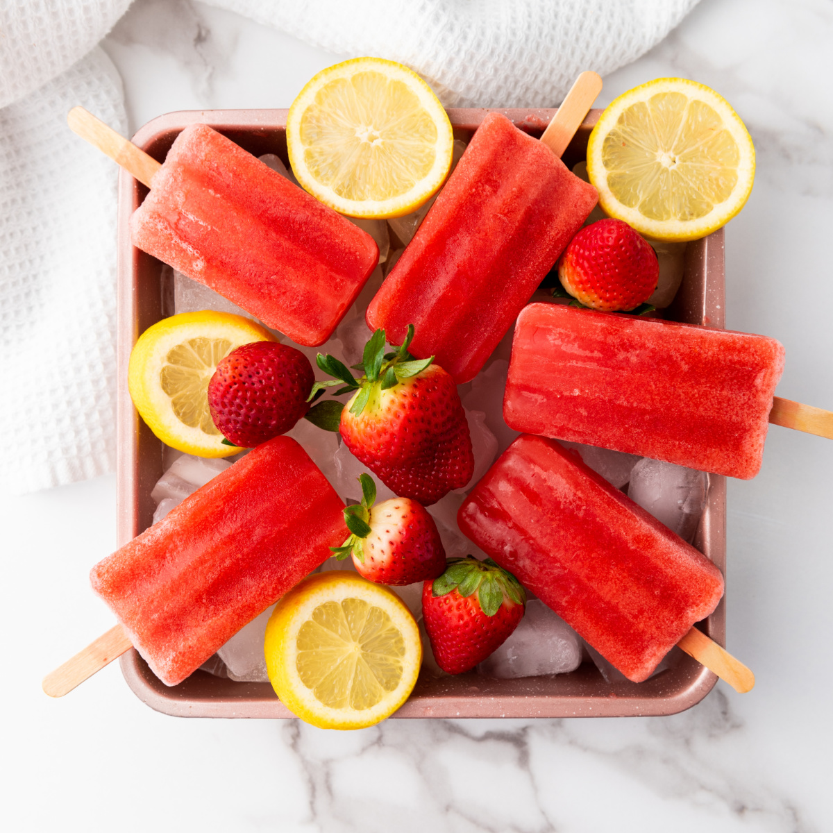 An overhead shot displays a pink tray of bright red strawberry lemon popsicles on sticks, nestled among ice cubes, whole fresh strawberries, and sliced lemons, all arranged on a white marble surface with a white towel in the upper left corner.
