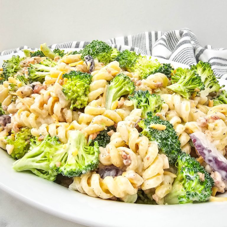 A wide shot of a white oval serving platter filled with creamy broccoli pasta salad, showcasing rotini pasta, green broccoli florets, and a creamy coating, set against a white background with a striped fabric detail.