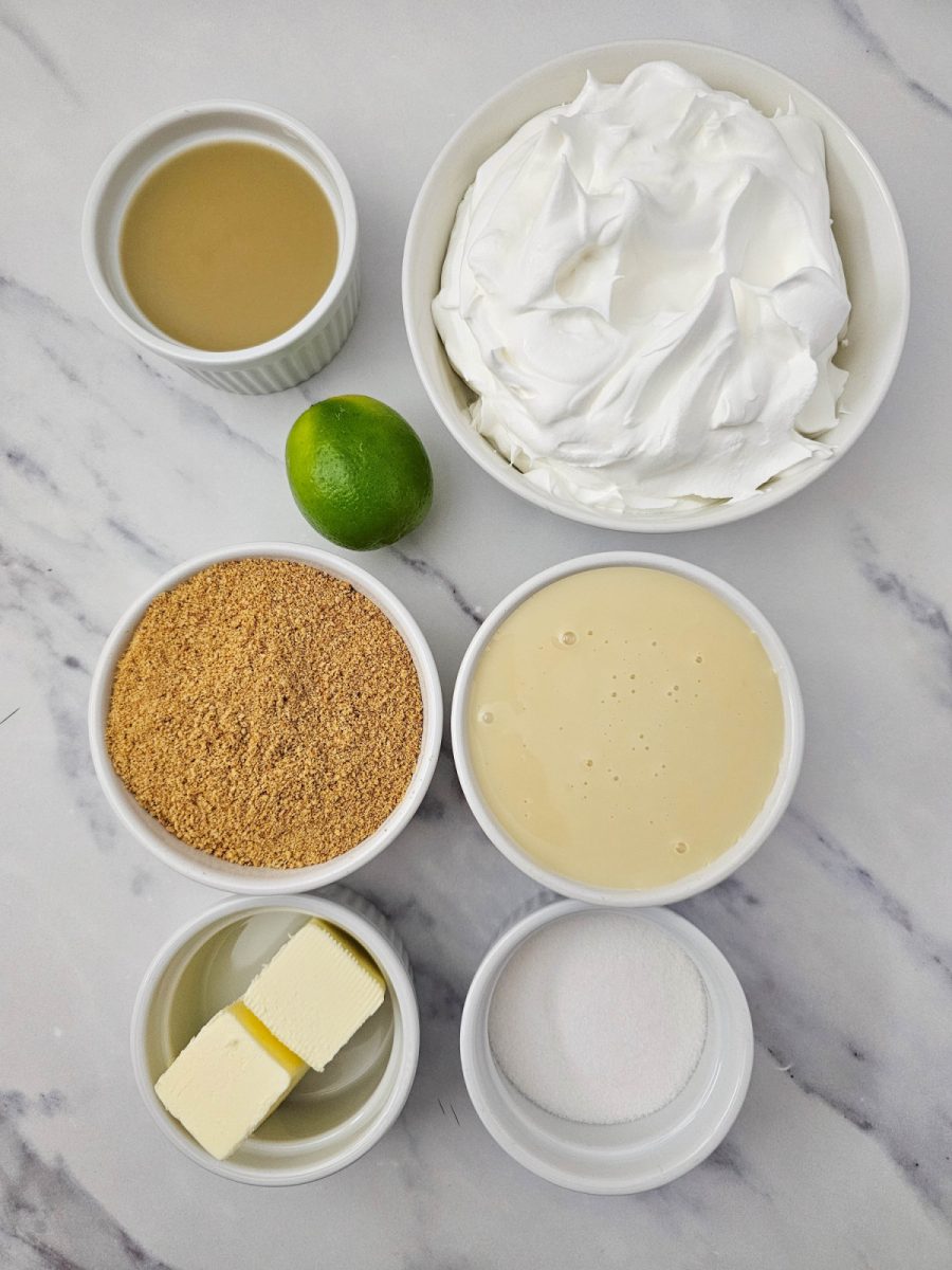Overhead view of white ramekins holding ingredients for key lime pie. Clockwise from top left: key lime juice, whipped topping, sweetened condensed milk, granulated sugar, melted butter, and graham cracker crumbs. A whole green lime sits in the center.