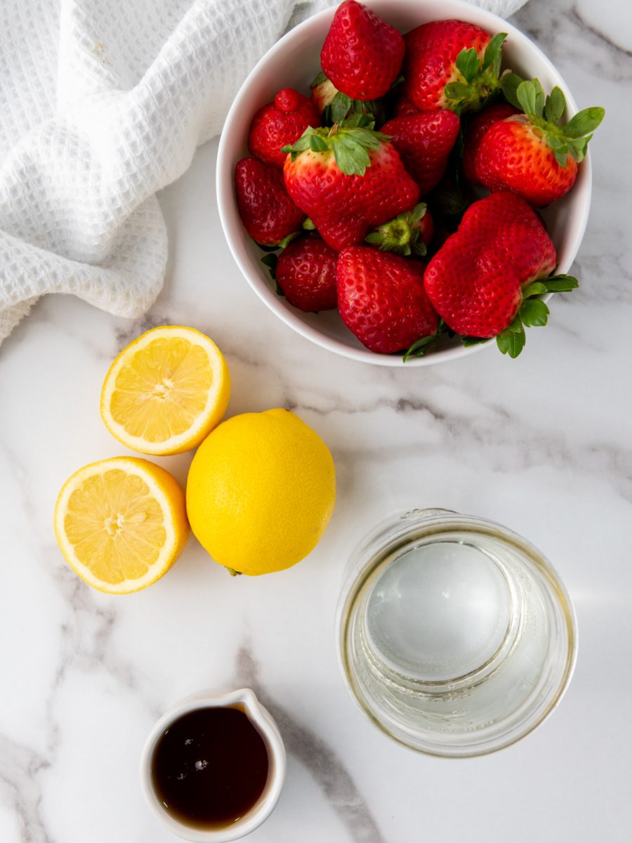  A white bowl filled with fresh, red strawberries sits on a white marble countertop next to two bright yellow lemons, a glass of water, and a small white dish containing a dark liquid, likely honey or maple syrup. A white kitchen towel is draped near the bowl of strawberries.