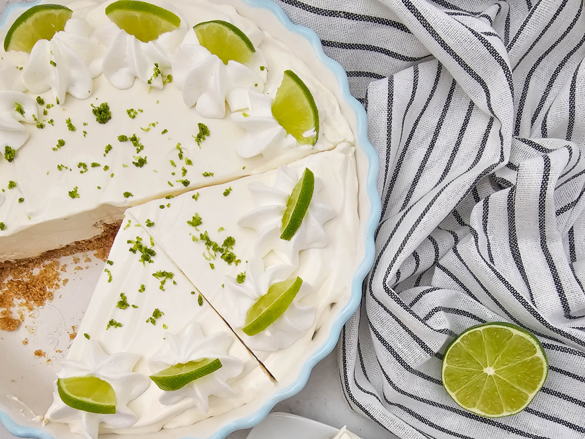 An overhead view of a whole easy key lime pie in a light blue pie dish, with one slice already cut out. The pie is topped with whipped topping, lime wedges, and green lime zest. A striped kitchen towel and whole lime are nearby.