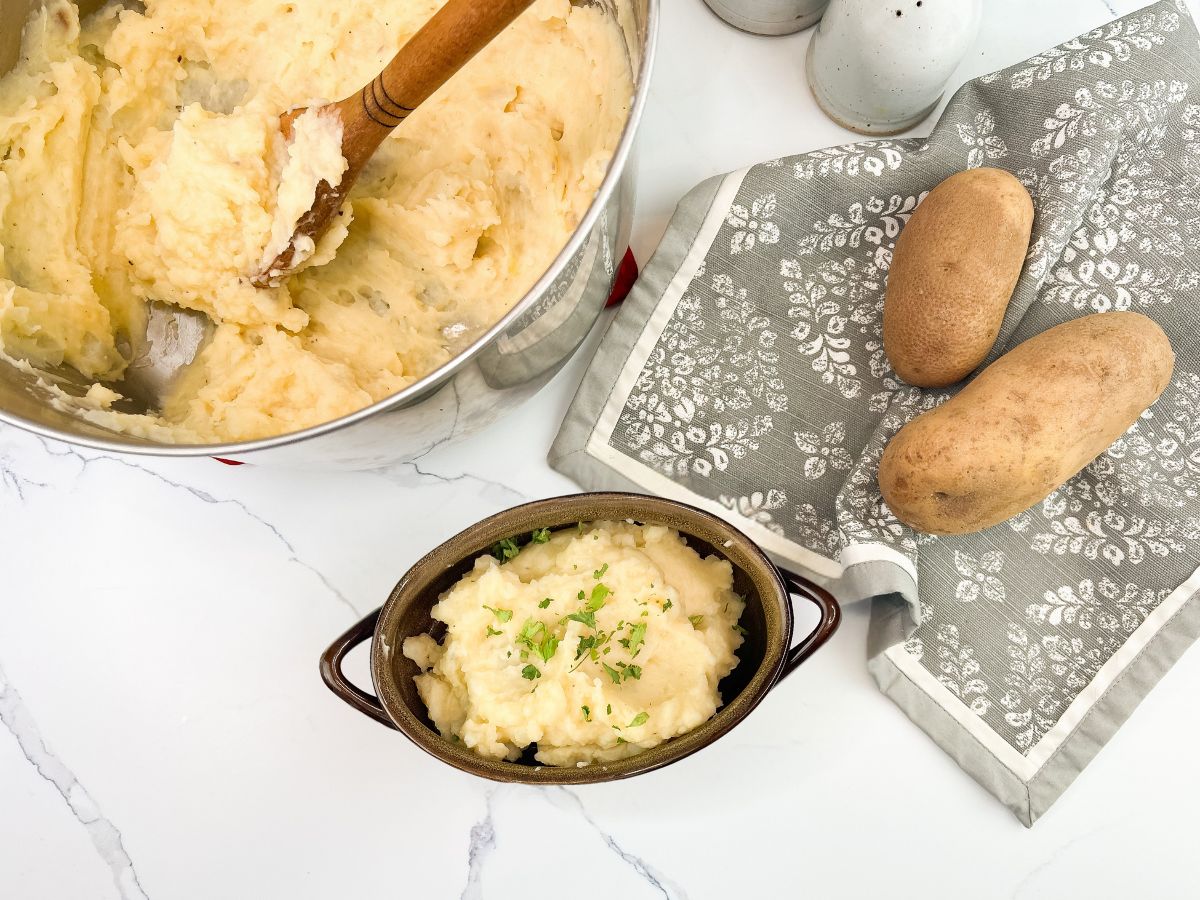 An overhead shot showing a large silver mixing bowl filled with creamy mashed potatoes and a wooden spoon resting inside. Below it, a smaller brown ceramic bowl holds a serving of mashed potatoes topped with parsley. To the right, two whole russet potatoes rest on a patterned grey and white kitchen towel, all on a white marble countertop.
