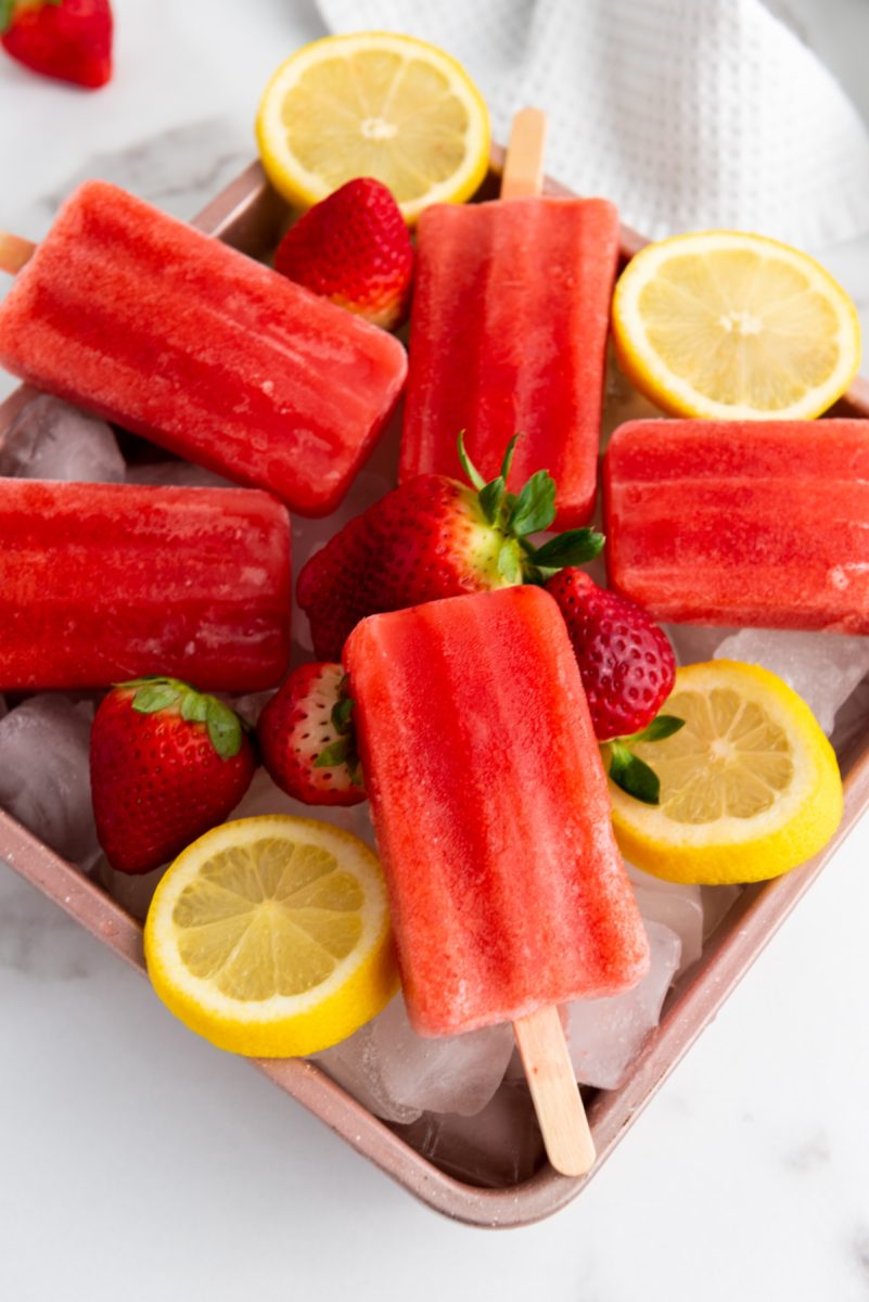 An overhead shot shows a pink tray filled with ice, holding multiple bright red fruit popsicles, fresh strawberries, and sliced lemons. A white kitchen towel is visible in the background on a marble surface.