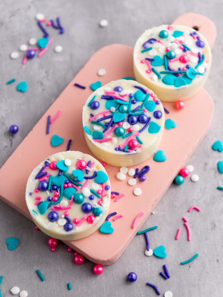 Overhead view of three round white chocolate covered Oreo cookies, each topped with a mix of pink, blue, and purple sprinkles, resting on a pink rectangular cutting board.