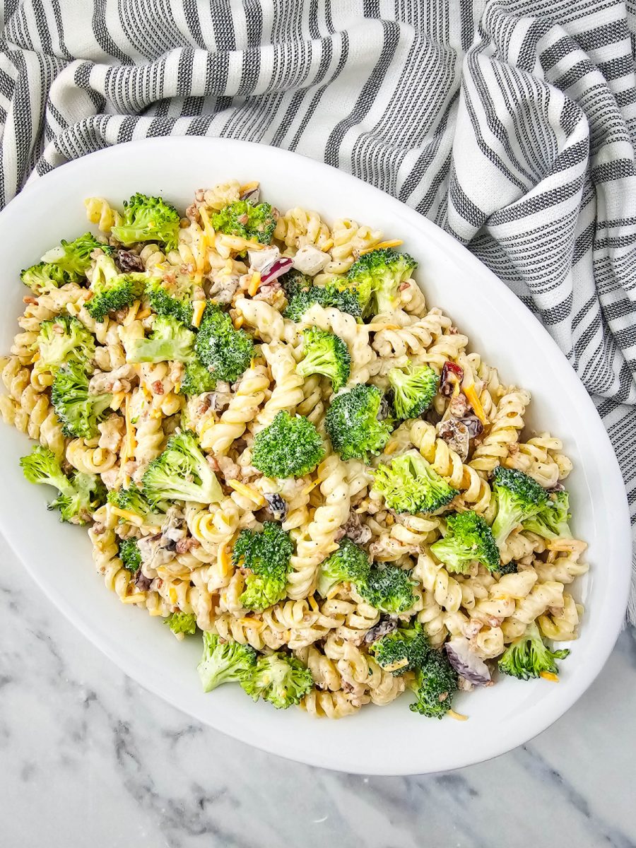 An overhead view of a creamy broccoli pasta salad served on a white oval platter, with a striped kitchen towel visible on the left side, presenting an arrangement of pasta, broccoli, and other ingredients.