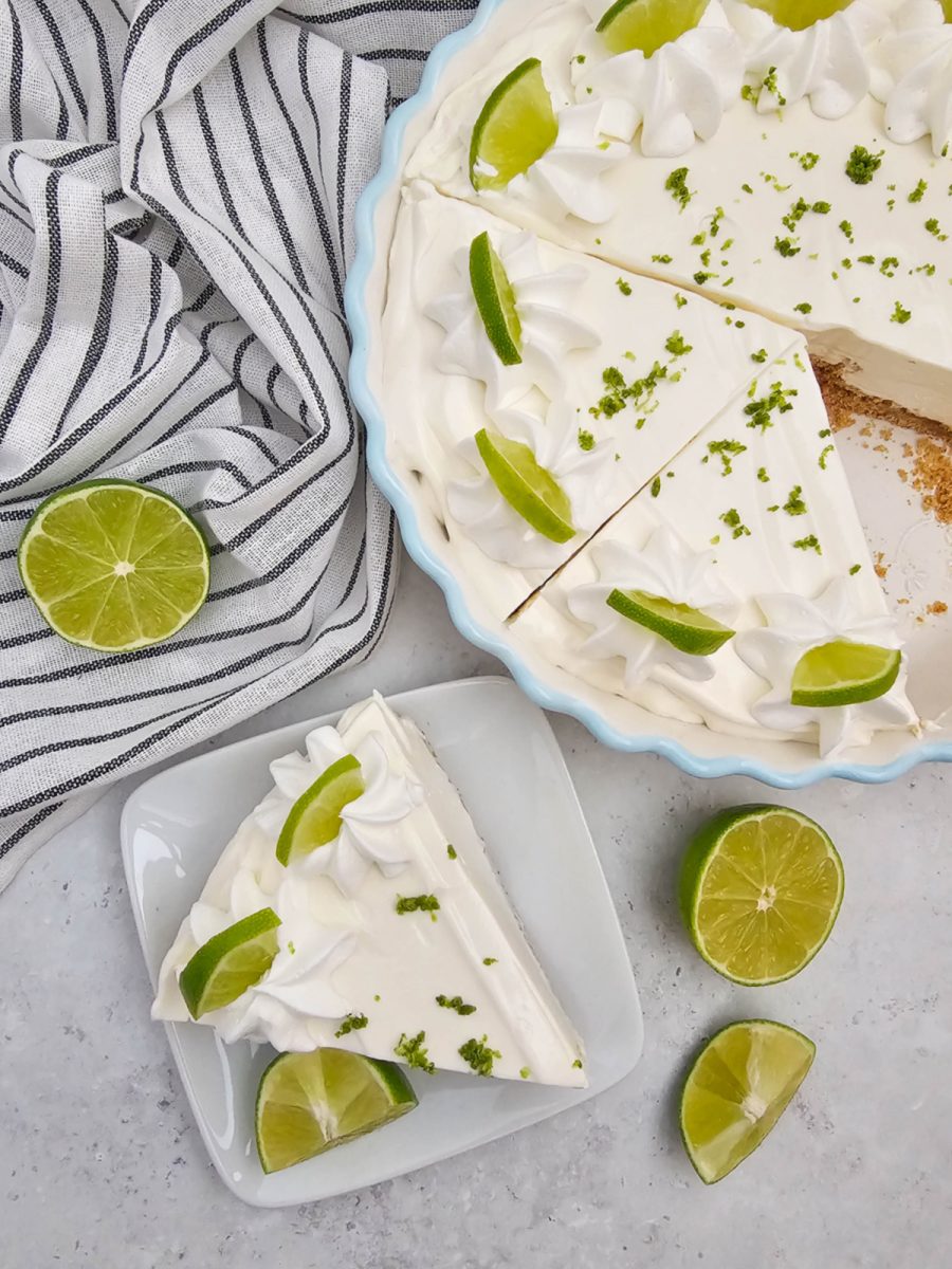 An overhead shot of a slice of easy key lime pie on a white square plate, adorned with whipped topping, lime wedges, and zest. In the background, the rest of the pie in a light blue dish shows where the slice was cut, with additional lime garnishes. A striped towel and lime halves are also visible.