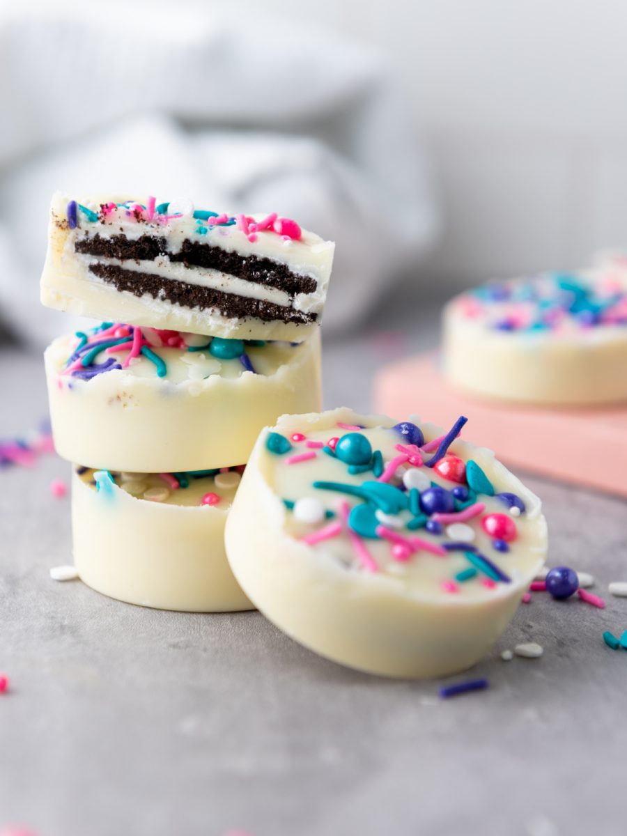 A stack of two white chocolate covered Oreo cookies topped with colorful sprinkles sits on a gray surface, with another white chocolate covered Oreo cookie in front. A white cloth and a pink board are in the background.