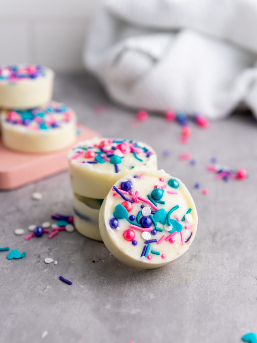  A stack of two white chocolate covered Oreo cookies topped with colorful sprinkles sits on a gray surface, with another white chocolate covered Oreo cookie in front. A white cloth and a pink board are in the background.