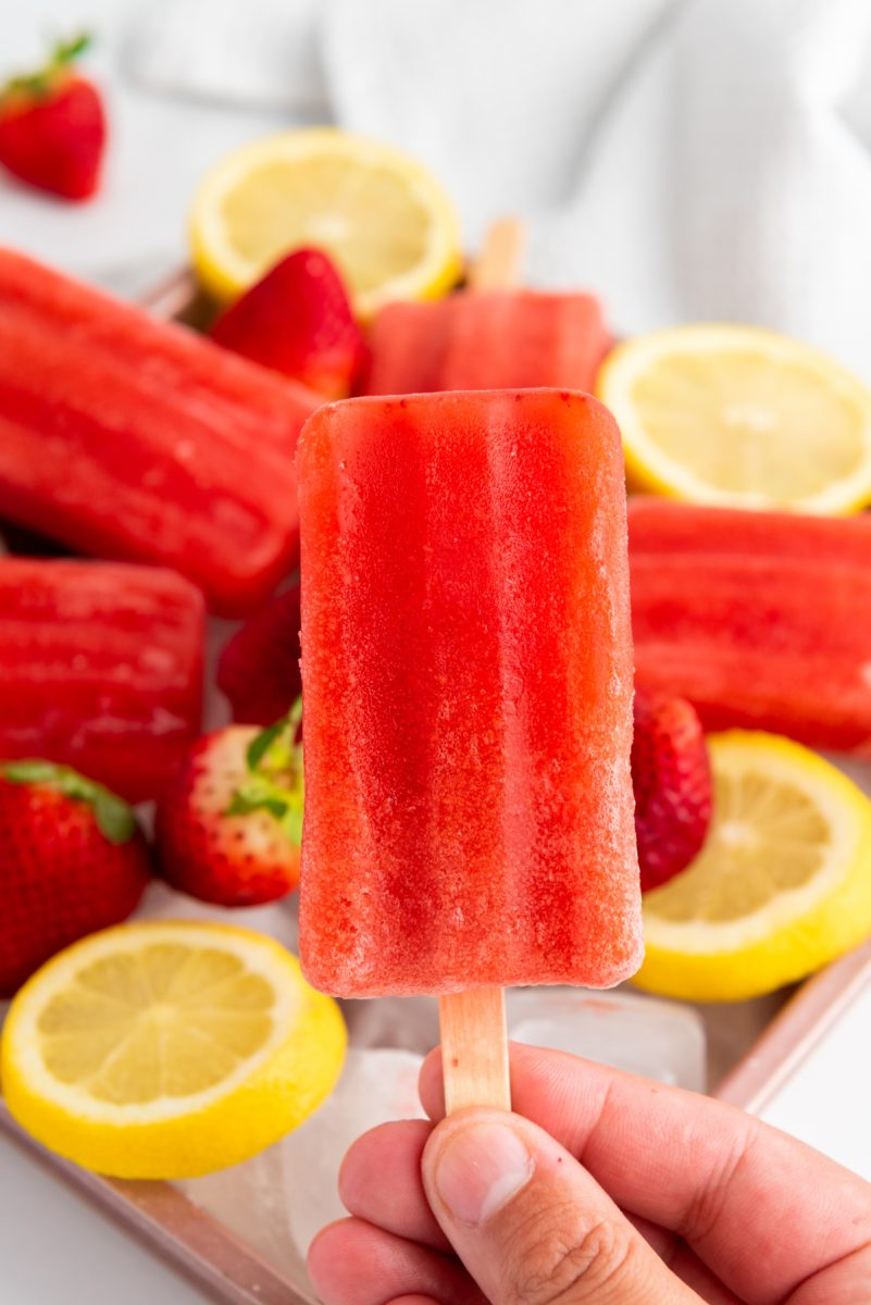 A hand holds up a single, bright red strawberry lemon popsicle against a blurry background of more popsicles, fresh strawberries, and lemon slices on ice. The popsicle shows a slightly frosted texture.